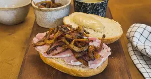 Open-faced ham sandwich with crispy fried onions and mustard on a cutting board, with coffee, napkins, and bowls in the background.