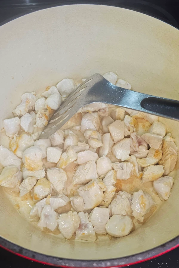 Cubed chicken breast lightly browning in a cream-colored Dutch oven with a slotted turner