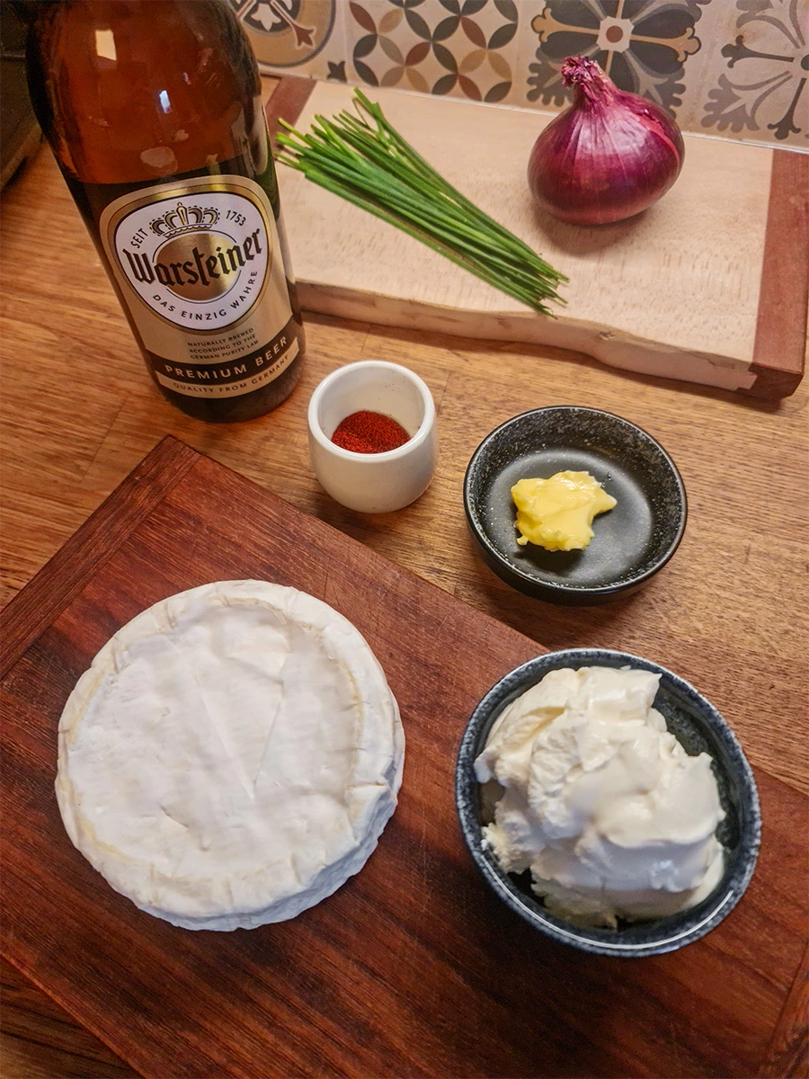 Ingredients for Obatzda including Camembert, cream cheese, butter, paprika, red onion, chives, and German beer.