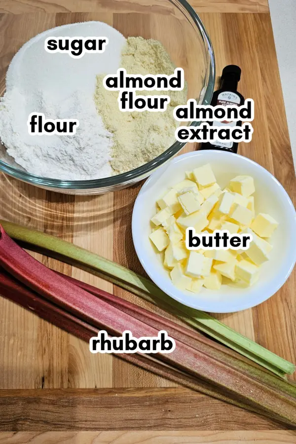 Labeled ingredients for rhubarb cake on a wooden surface, including flour, sugar, almond flour, almond extract, butter, and fresh rhubarb stalks.