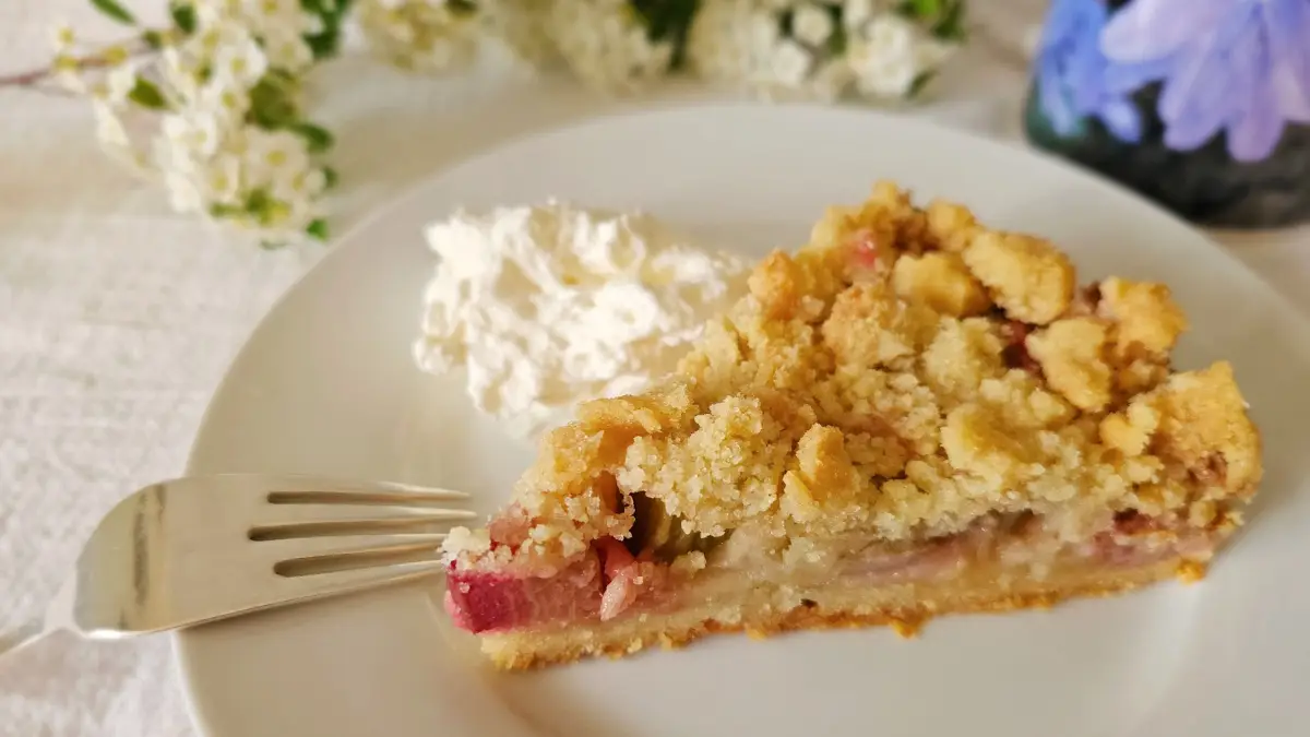 Slice of rhubarb cake with golden streusel topping, served on a white plate with a dollop of whipped cream and a fork beside it.