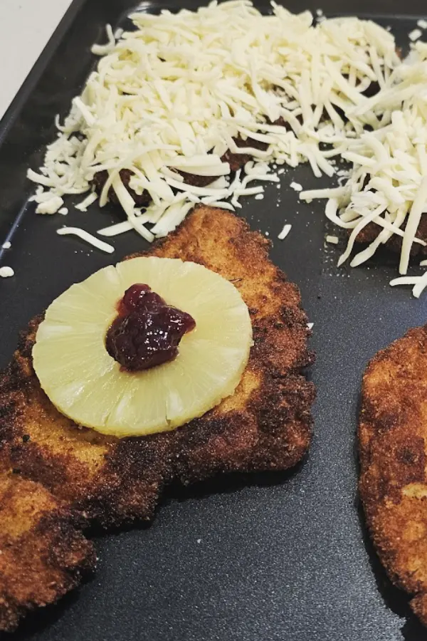 Fried chicken schnitzel topped with a canned pineapple ring and cranberry sauce on a baking sheet