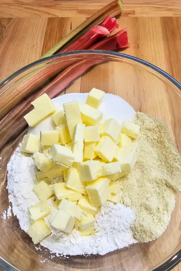 Glass bowl filled with cubed butter, flour, sugar, and almond flour, with fresh rhubarb stalks in the background on a wooden surface.