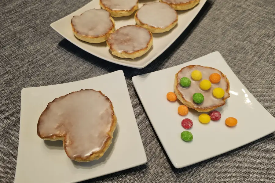 Heart-shaped Amerikaner and an iced Amerikaner decorated with colorful candies on white plates, with more German iced cookies behind