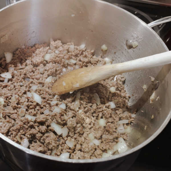 Ground beef and chopped onions browning in a stainless steel pot with a wooden spoon.