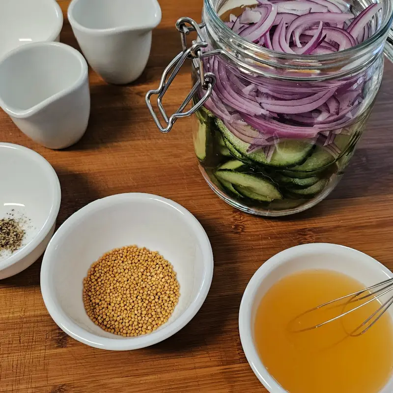 Sliced cucumbers and red onion layered in a glass jar beside mustard seeds and mixed vinegar dressing