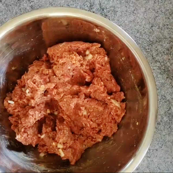 Ground beef mixture with onions and seasonings combined in a stainless steel bowl.
