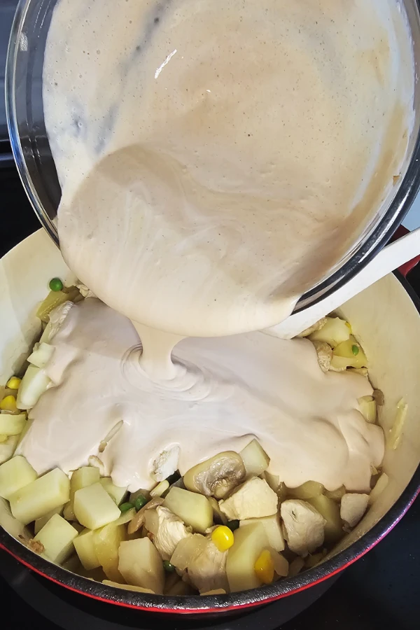 Creamy curry sauce poured from a glass bowl over chicken, potatoes, corn and peas in a Dutch oven