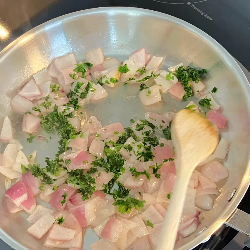 Chopped onion and parsley sautéing in a skillet with a wooden spoon.