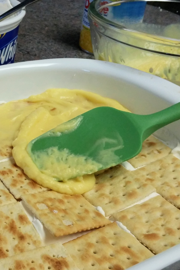 Vanilla pudding being spread over a layer of soda crackers in a white dish.