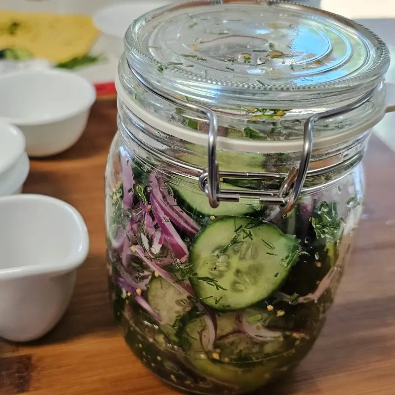 Covered glass jar filled with cucumber slices, red onion, dill, mustard seeds, and vinegar dressing