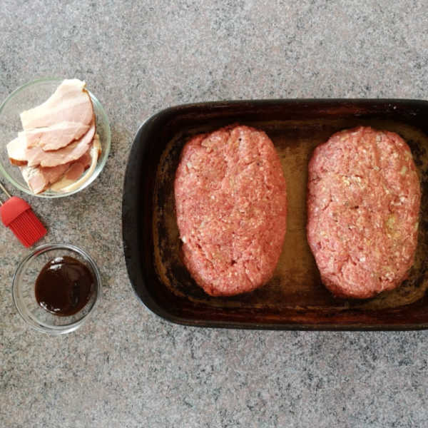 Two raw meatloaf loaves shaped in a baking dish, with bacon and BBQ sauce on the side.