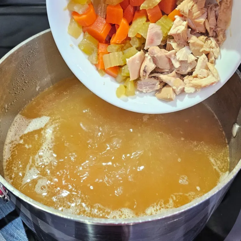 Adding cooked chicken and vegetables to simmering broth to extend German egg drop soup (Einlaufsuppe).