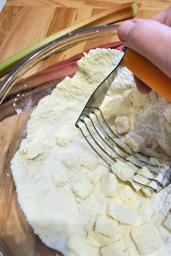Hand using a pastry cutter to blend cubed butter into flour mixture in a glass bowl, with rhubarb stalks nearby on a wooden surface.
