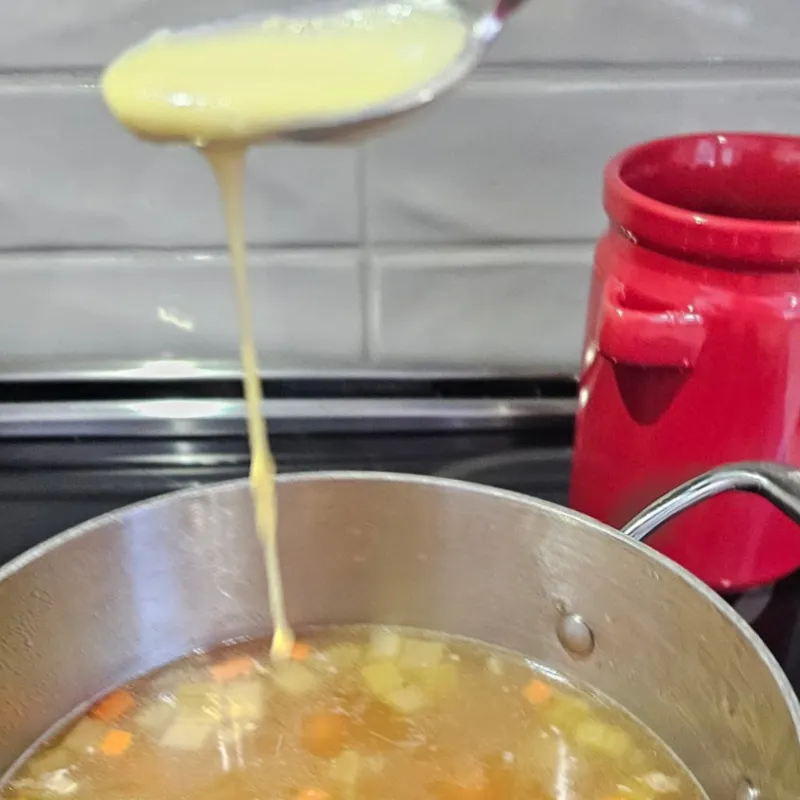 Dripping egg and flour batter into simmering broth while stirring for German egg drop soup (Einlaufsuppe).