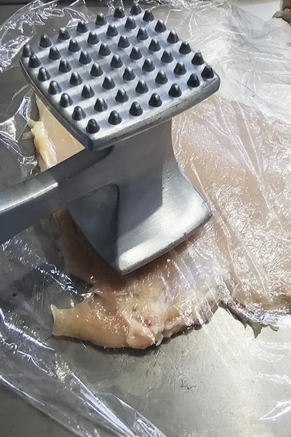 Meat mallet pounding a chicken breast cutlet between plastic wrap to an even thickness