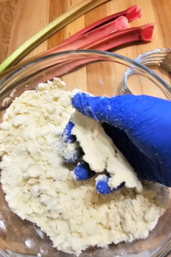 Gloved hand crumbling streusel mixture in a glass bowl with fresh rhubarb stalks in the background.