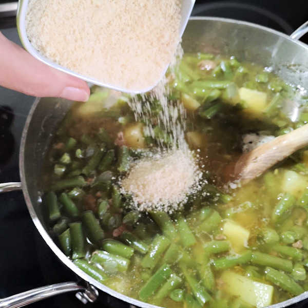 Instant mashed potato flakes being sprinkled into a pot of simmering green bean soup.