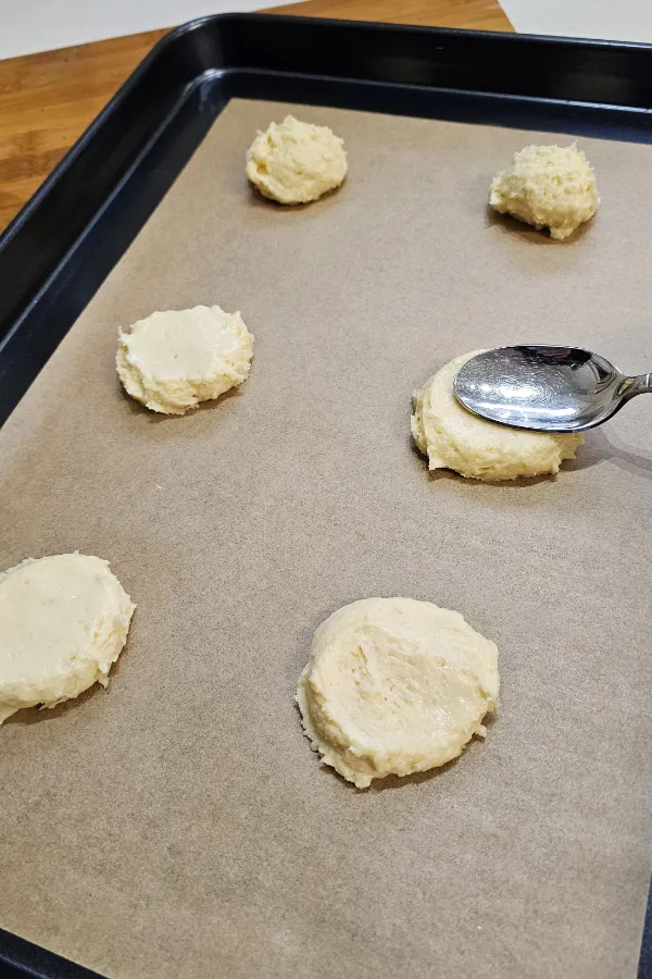Spoonfuls of Amerikaner batter on a parchment-lined baking sheet, with a spoon smoothing one mound