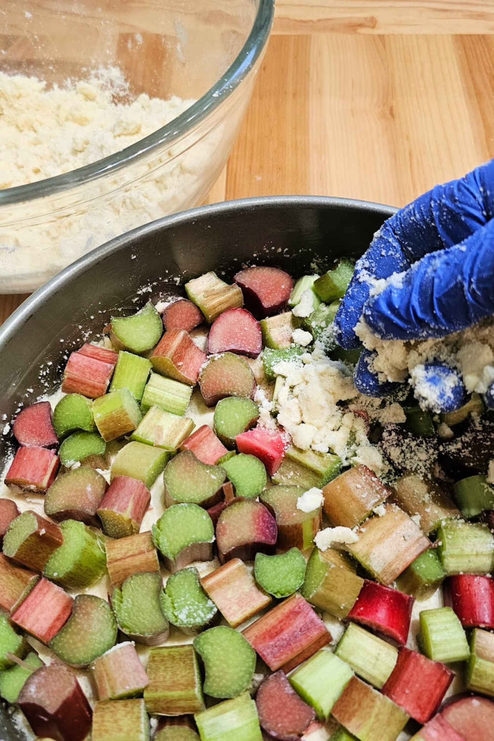 Fresh chopped rhubarb in a cake pan being topped with streusel mixture by a gloved hand, with a mixing bowl in the background.