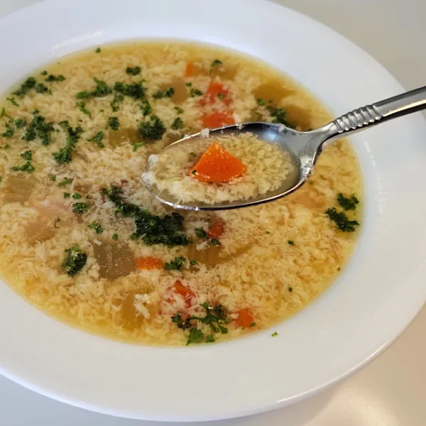 Finished German egg drop soup (Einlaufsuppe) in a bowl with egg wisps, carrots, herbs, and broth on a spoon.