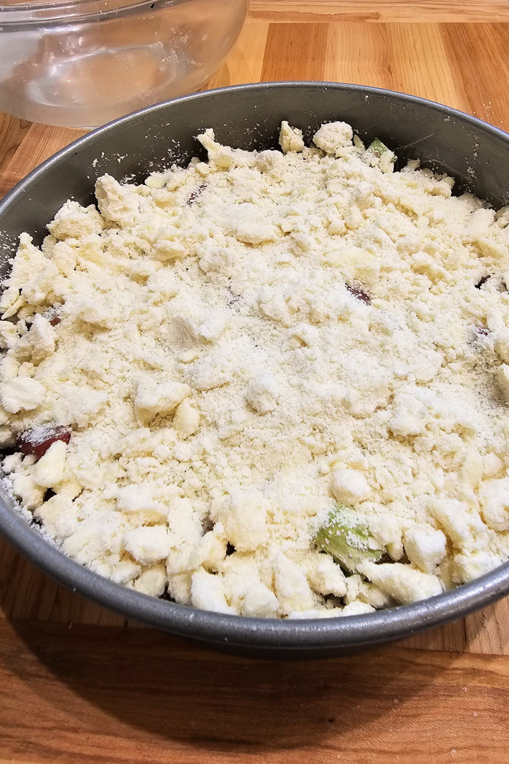 Round baking pan filled with unbaked rhubarb streusel cake, topped generously with raw crumble topping, ready to go in the oven.
