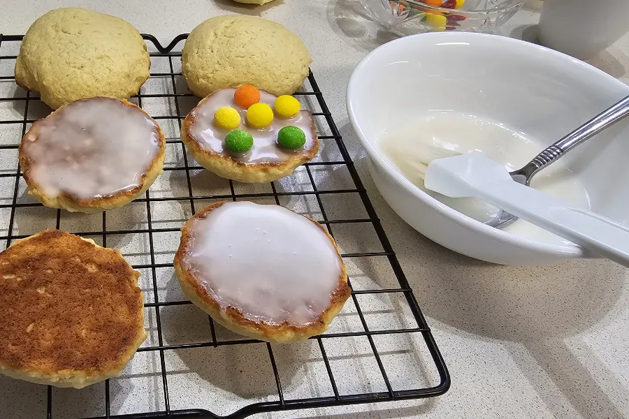 Amerikaner cookies on a wire rack, some iced on the flat side and one topped with candies, next to a bowl of white icing