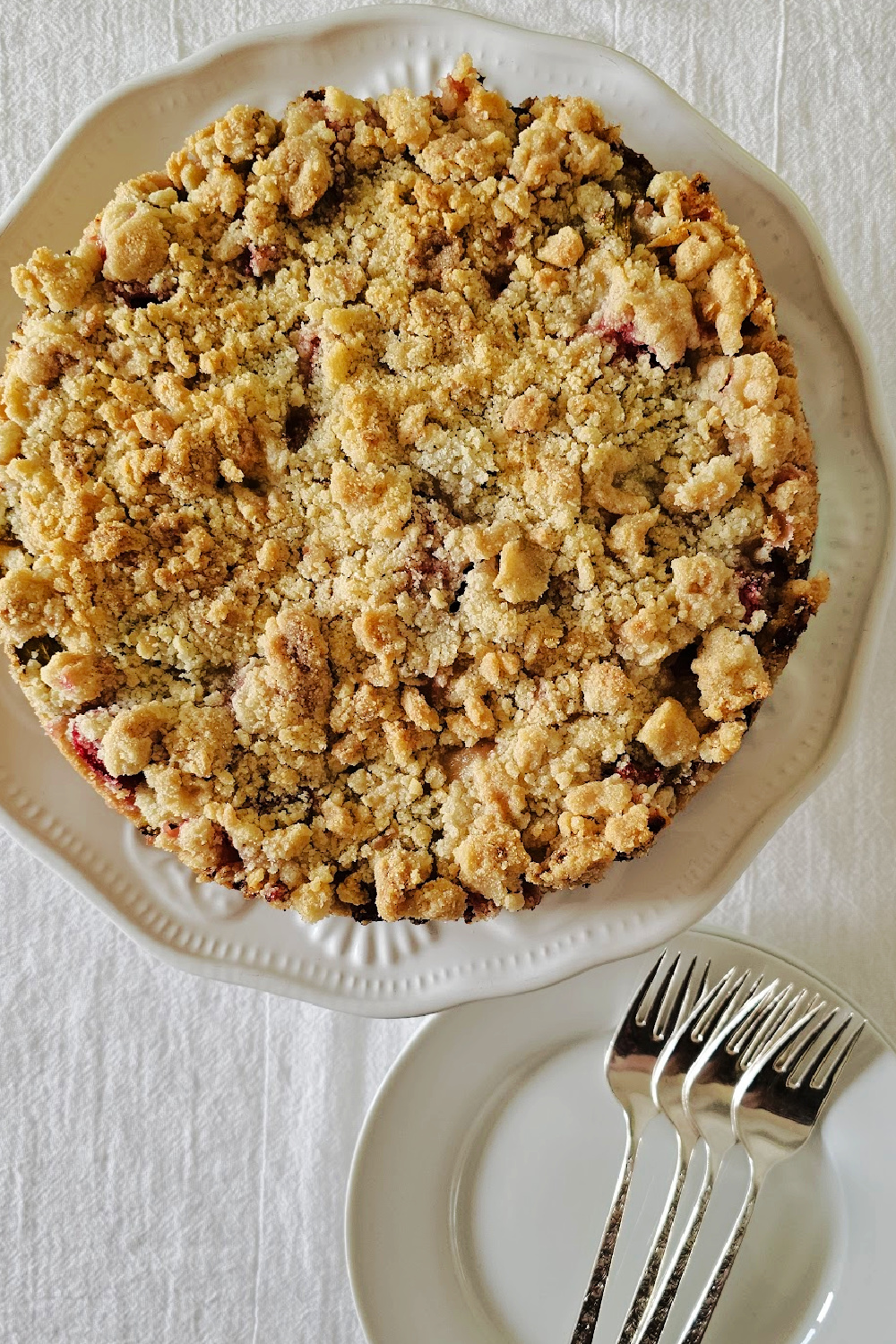 Whole rhubarb streusel cake with golden topping on a white platter, beside a plate with forks, ready to serve.