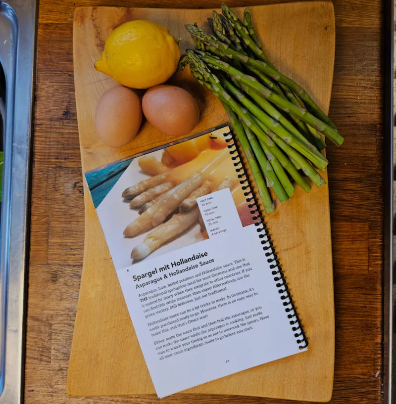 Open spiral-bound Easter Feast cookbook to Spargel mit Hollandaise with fresh asparagus, eggs, and a lemon on a cutting board