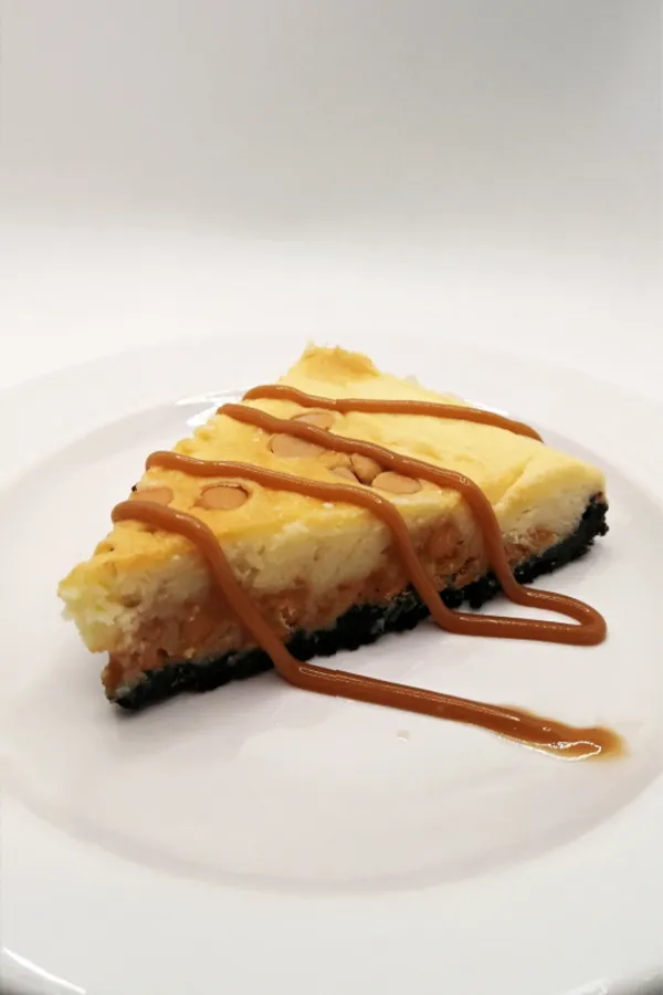 A slice of cheesecake sits on a white plate, drizzled with caramel sauce and featuring a dark crumbly crust.