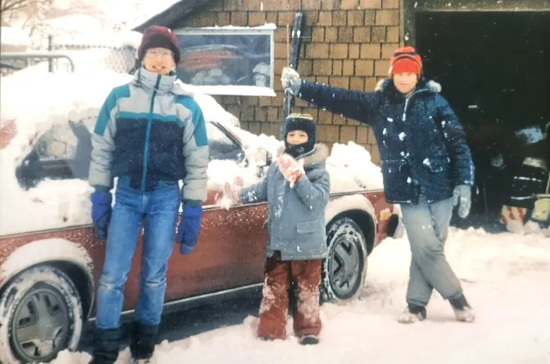 Young Andrew (left) stands in the snow with his brothers Eran (middle) and Mike (right) beside a snow-covered car during a Canadian winter, bundled up in winter coats, hats, and gloves.