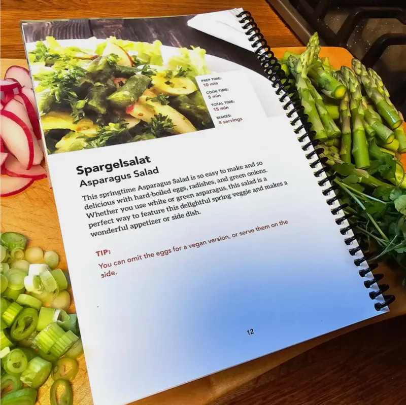 Open spiral-bound Easter Feast cookbook to the Spargelsalat (Asparagus Salad) page on a cutting board with fresh asparagus, radishes, and chopped green onions