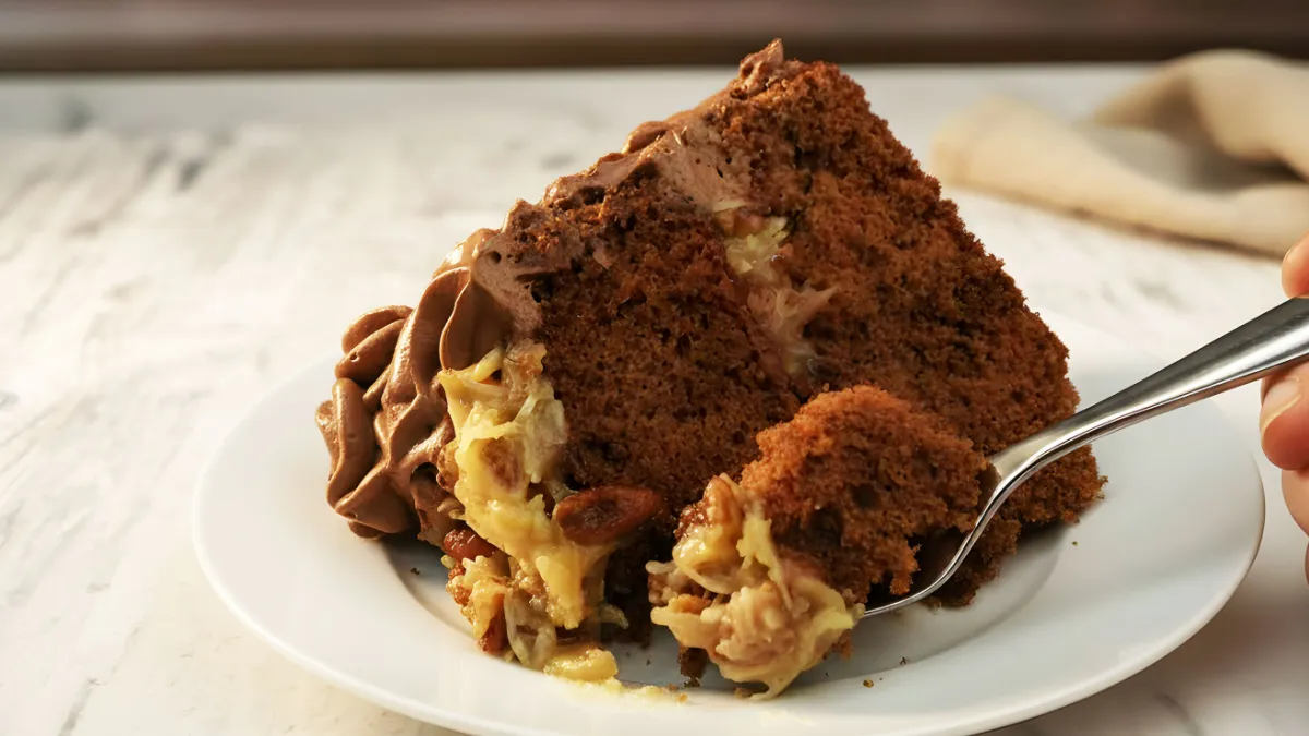 Slice of German chocolate cake on a white plate with coconut-pecan filling and chocolate frosting, with a forkful taken from the front.