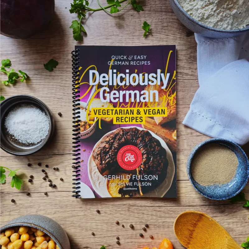 Spiral-bound Deliciously German Vegetarian & Vegan Recipes cookbook on a wooden table with bowls of flour, salt, yeast, herbs, and chickpeas