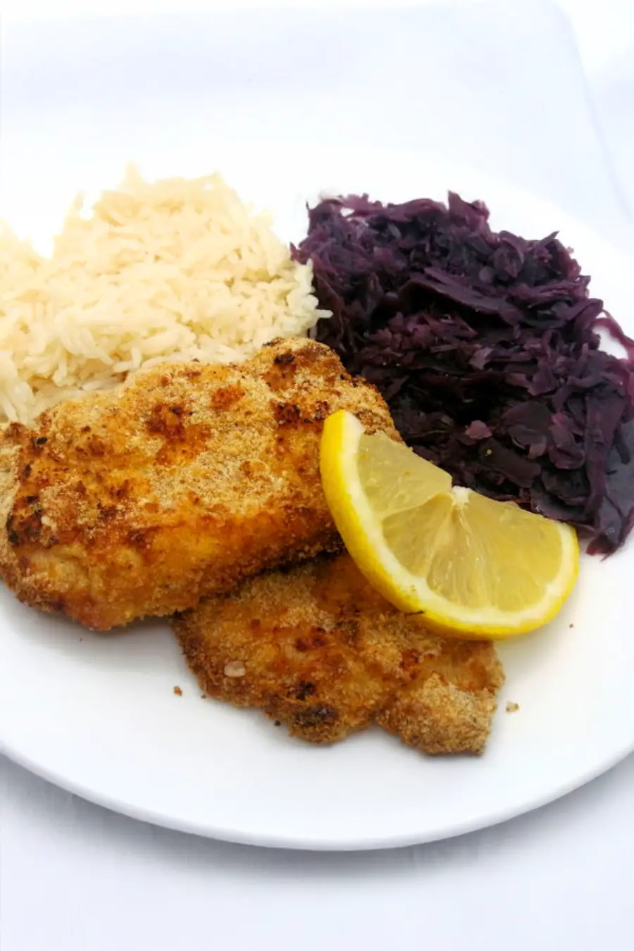 A plate with schnitzel, two lemon slices, a serving of white rice, and a portion of cooked red cabbage.