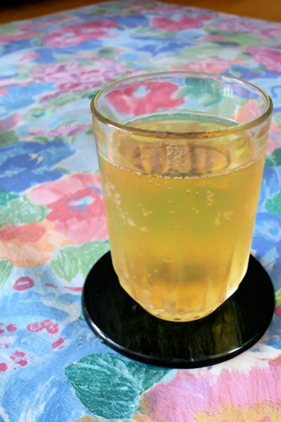 A glass of sparkling apple spritzer sits on a black coaster, placed on a floral tablecloth.