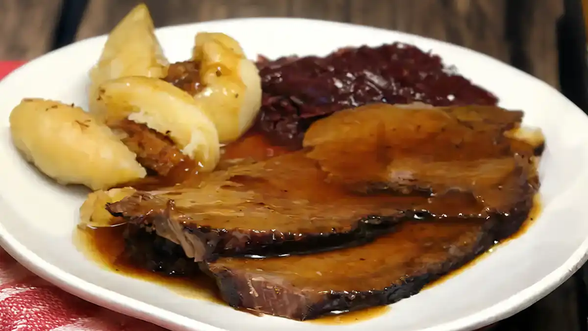 White plate with slices of sauerbraten (marinated beef) with gravy. Potato dumplings and red cabbage are the sides to the meat.