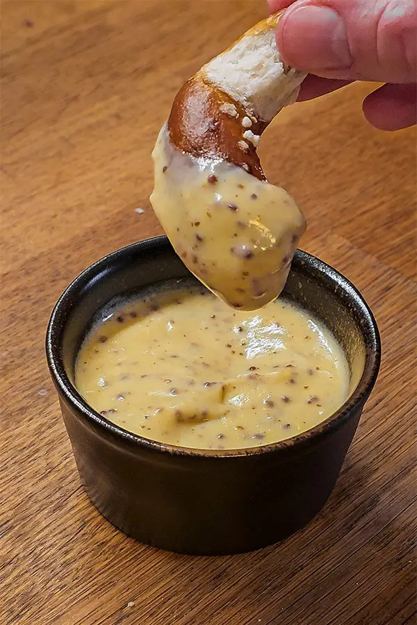 Thick and creamy beer mustard sauce in a dark ramekin, with a hand dipping a Bavarian pretzel twist into the sauce.