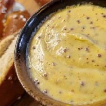 Close-up of creamy beer mustard sauce with visible mustard seeds in a rustic ceramic bowl, served alongside soft German pretzels.
