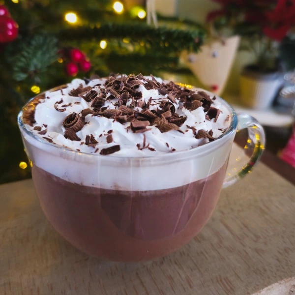 Glass mug of Black Forest hot chocolate topped with whipped cream and chocolate shavings in front of a Christmas tree.