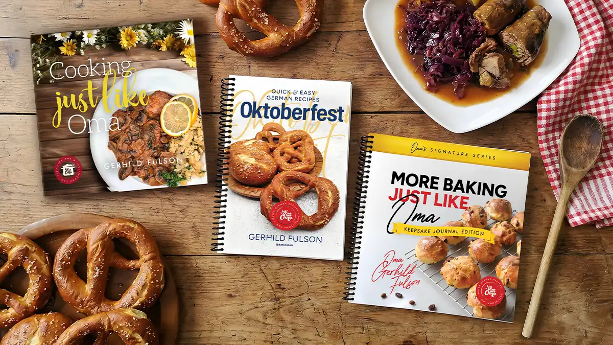 Three Just Like Oma German cookbooks in English by Gerhild Fulson with soft pretzels and a plate of rouladen and Rotkohl on a wooden table.
