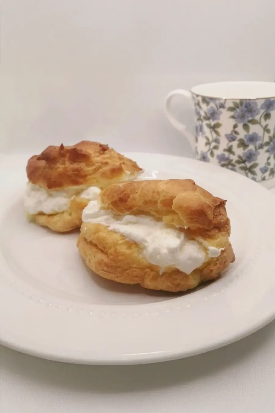 Two cream puffs on a white plate with a floral-patterned teacup in the background.