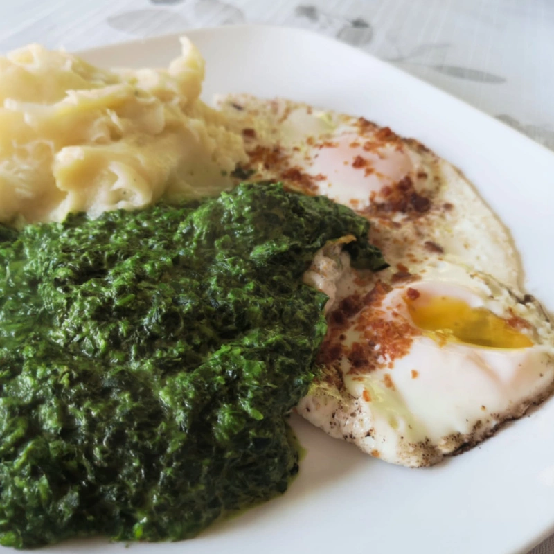 Plate of creamed spinach served with mashed potatoes and two fried eggs, showing the creamy texture and vibrant green color of the spinach.