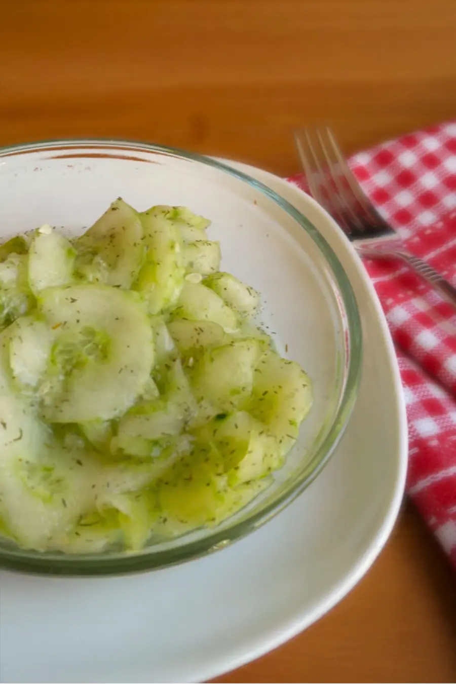 A glass bowl of cucumber salad with dill sits on a white plate. A red and white checkered cloth and a fork are on the wooden table beside it.