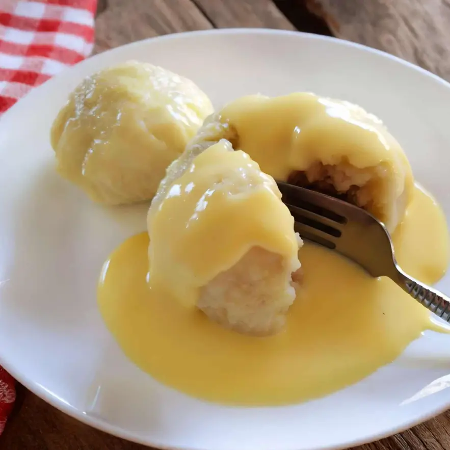 Two golden-brown Dampfnudeln on a white plate, one cut open to show the soft, fluffy inside, topped with creamy vanilla sauce