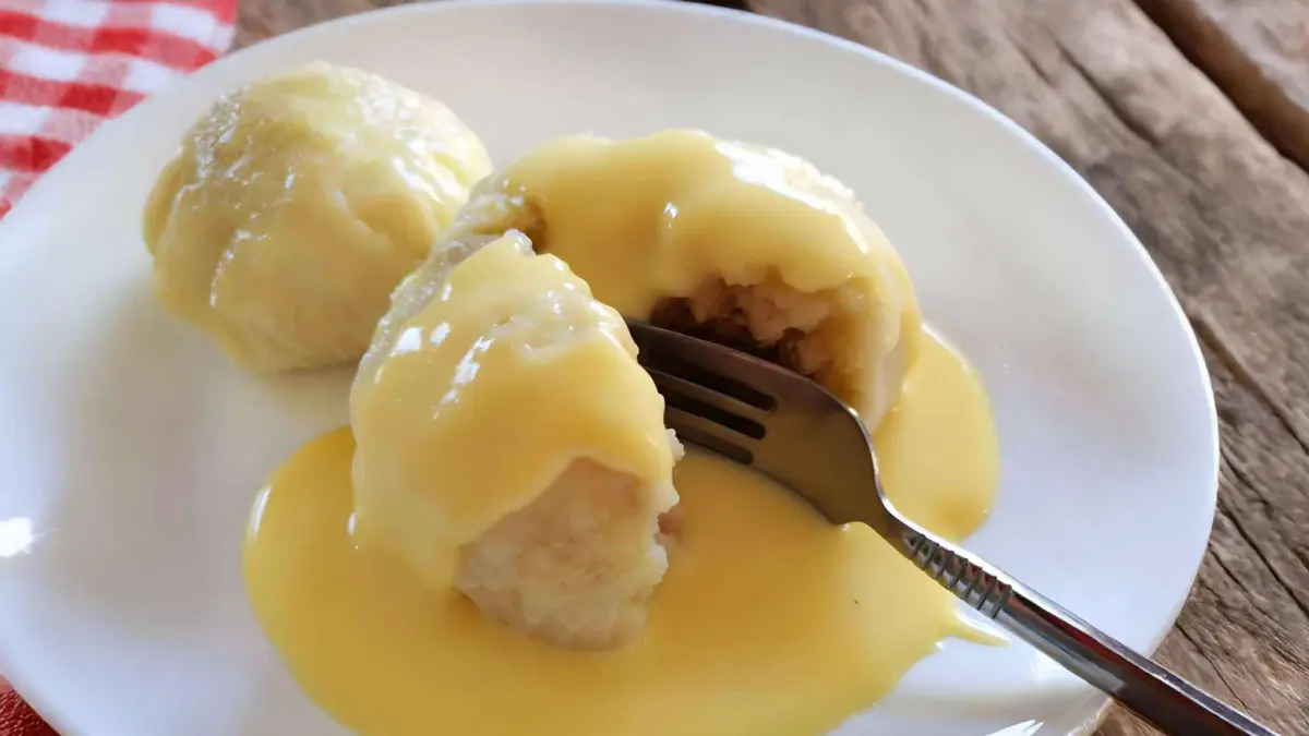 Dampfnudeln with Vanilla Sauce – Classic German Steamed Sweet Dumplings Two golden-brown Dampfnudeln on a white plate, one cut open to show the soft, fluffy inside, topped with creamy vanilla sauce.