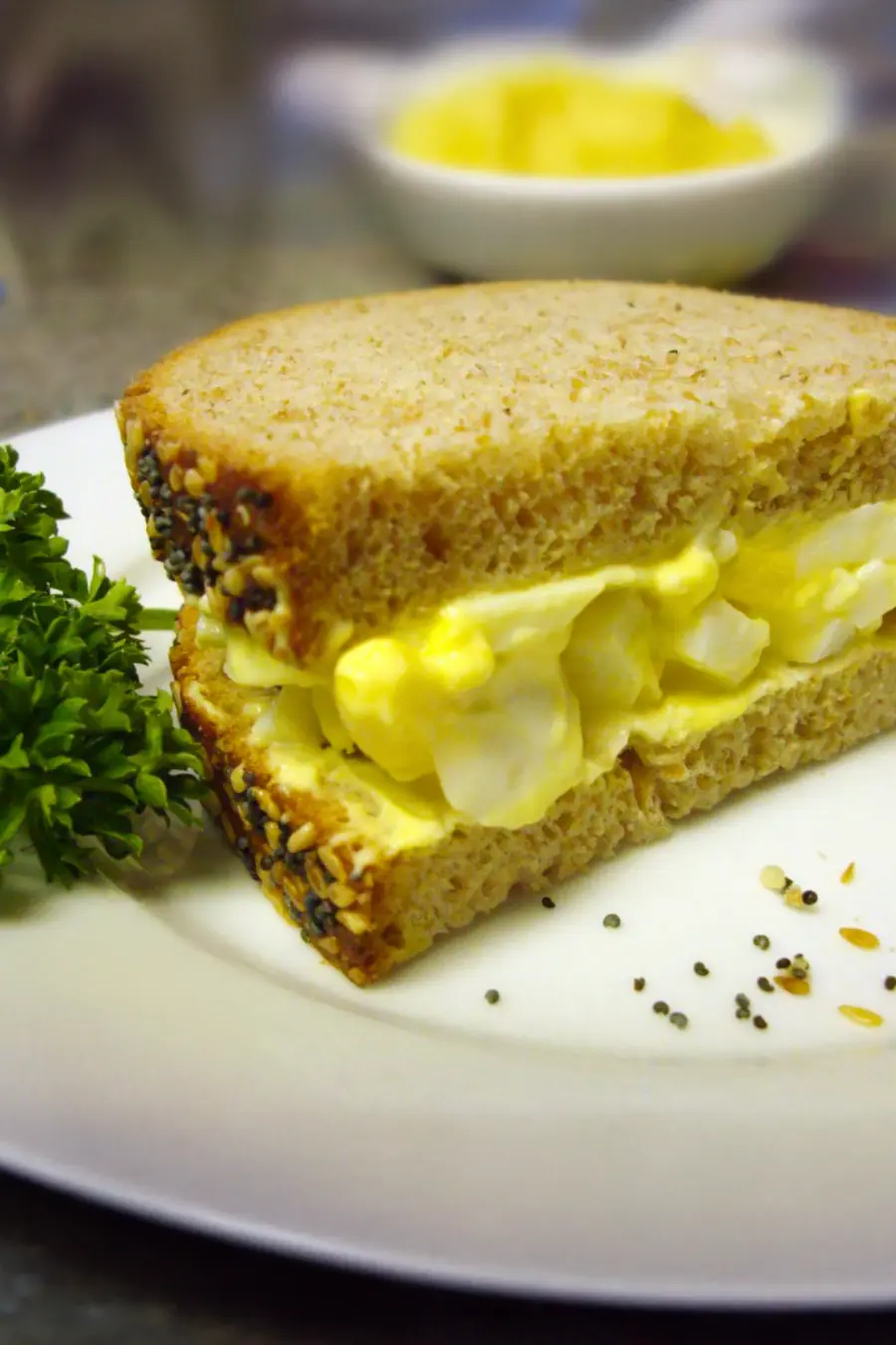 Egg salad sandwich on whole grain bread, garnished with parsley on a white plate.