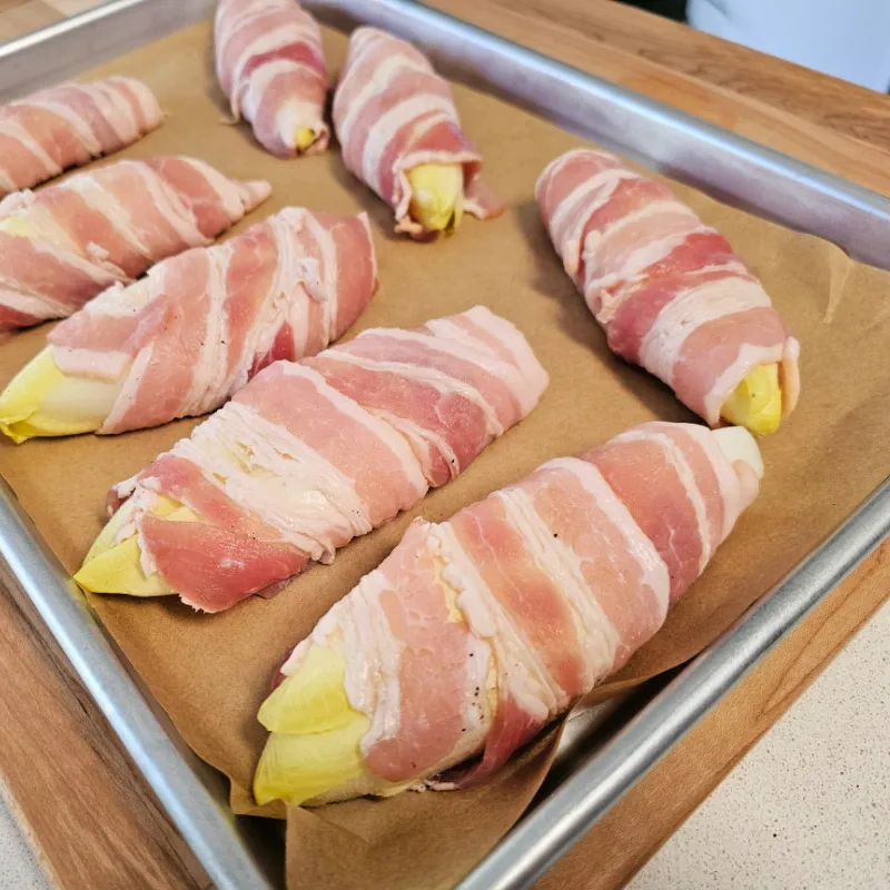 Belgian endive halves wrapped in bacon on a parchment-lined baking sheet before baking.