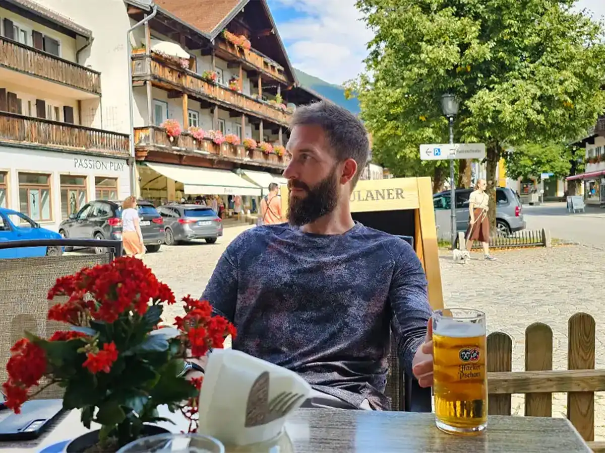 Eran Fulson enjoying a beer in a Bavarian village, surrounded by traditional Alpine buildings and flower boxes.