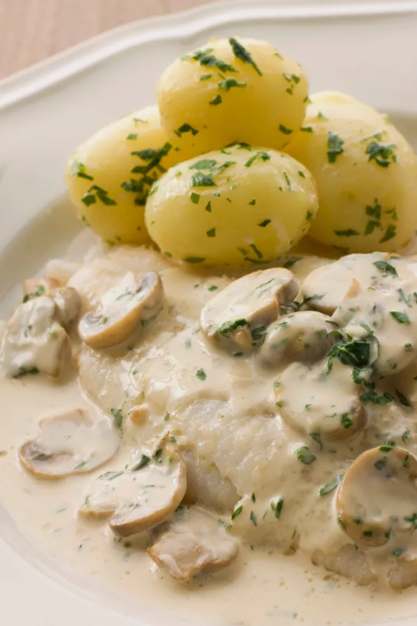 German fricasseed fish in creamy mushroom sauce, served with parsley potatoes on a white plate.